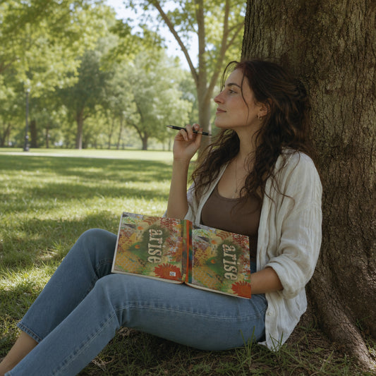 Woman sitting under a tree in a park, reflective journaling in her Arise and Be Seen hardback journal.
