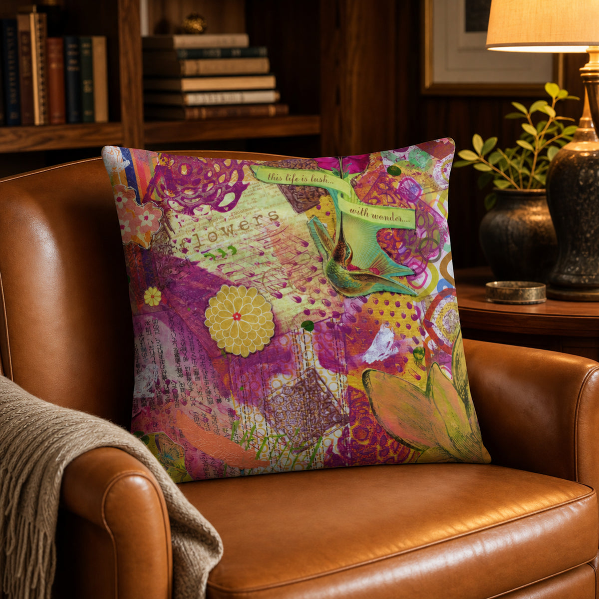An 18x18" square decorative throw pillow with a colorful, artistic floral and botanical print is styled on a brown leather armchair in a cozy, warmly lit living room setting.