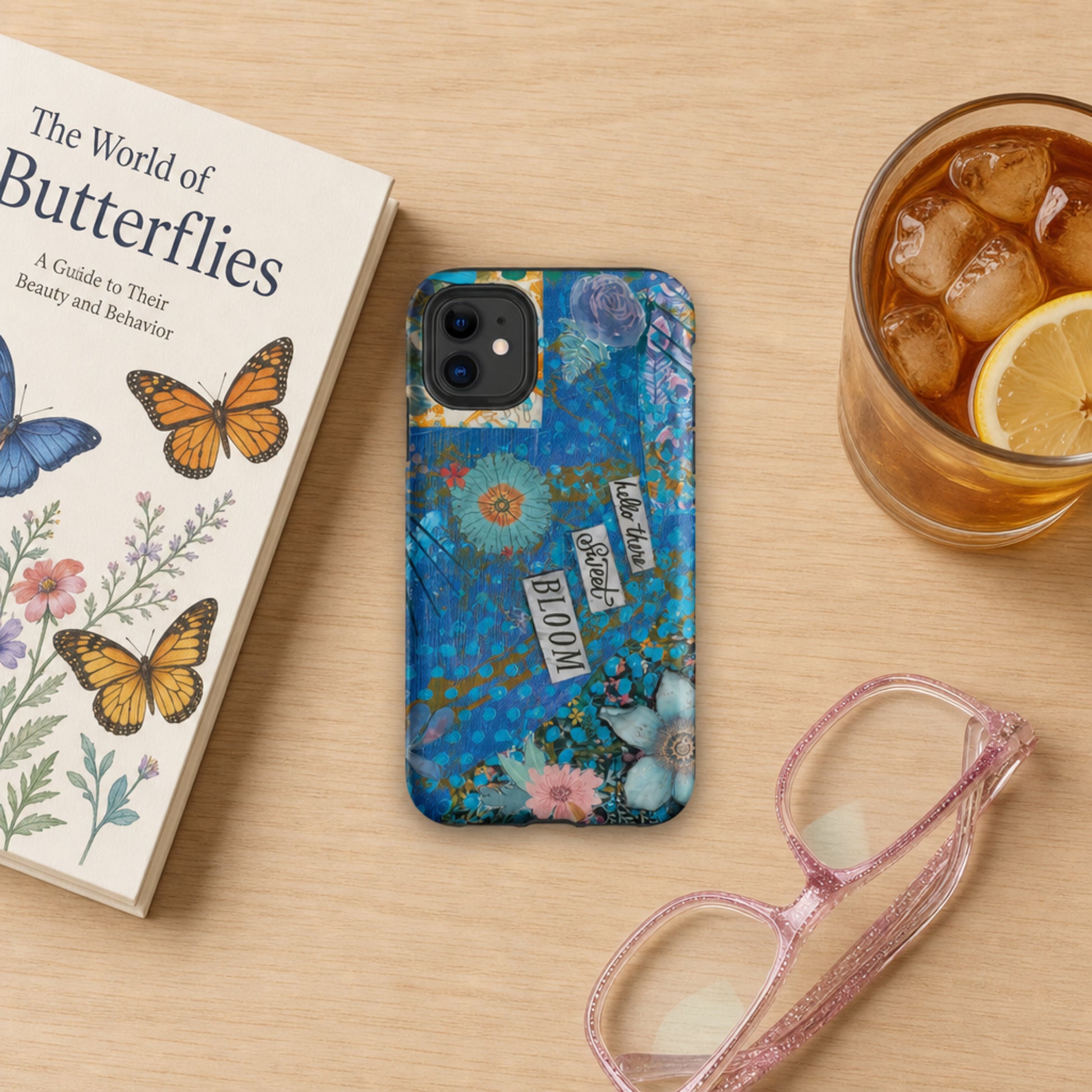 An image of the blue floral phone case with hello there sweet bloom quote laying on a wooden table with a book, eyeglasses and a glass of iced tea near it.