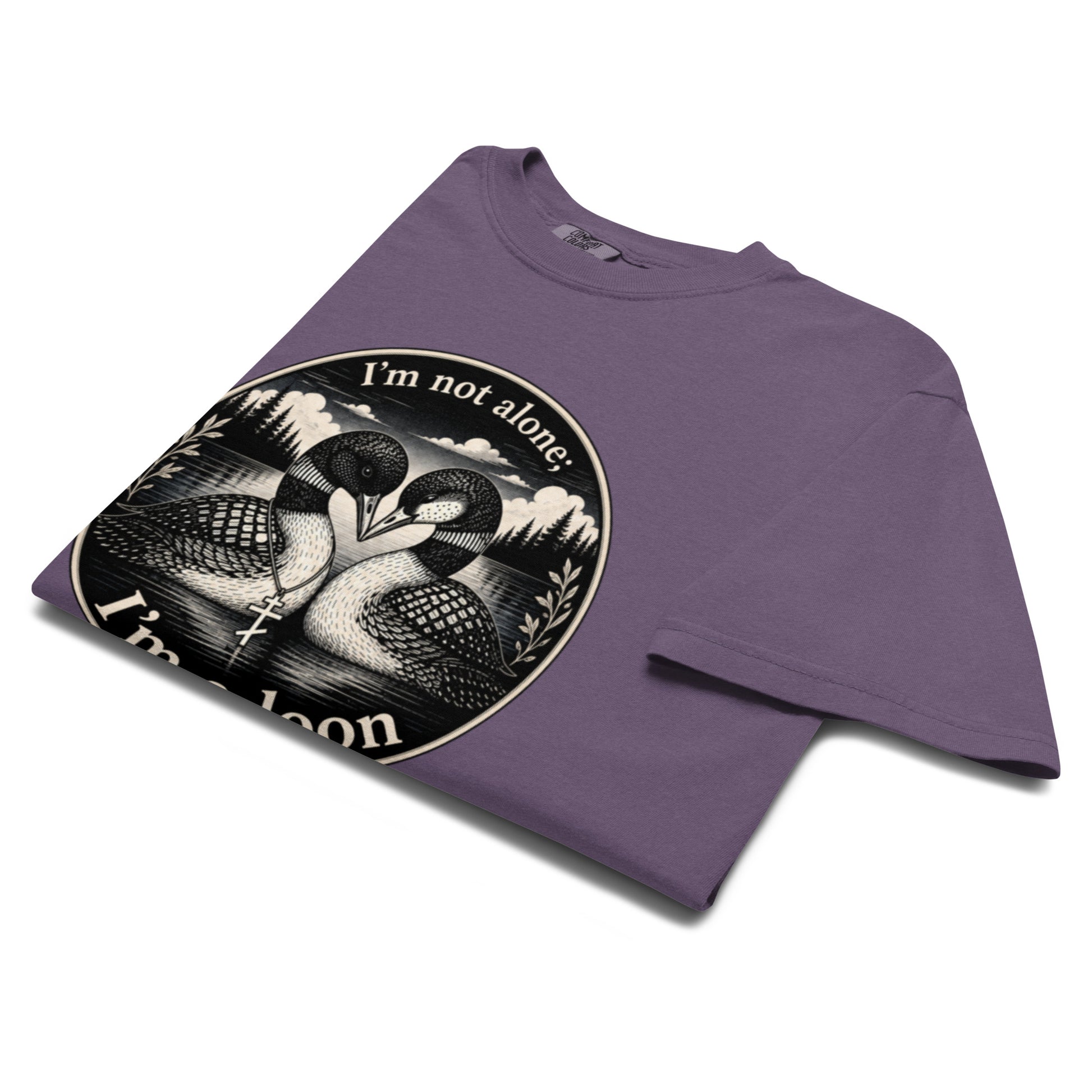 A folded grape colored crewneck t-shirt with a circular black and white graphic print of two loons and the text 'I'm not alone' and 'I'm a loon'.